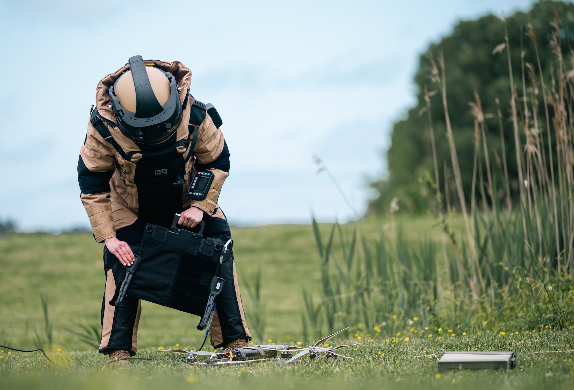A woman in a bomb suit inspects a drone in the grass