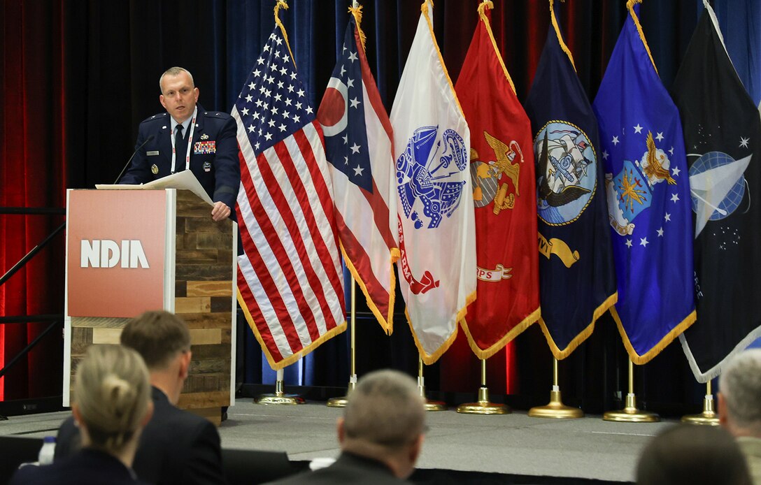 A man in a blue Air Force uniform speaks at a podium with a backdrop of flags in a large ballroom.
