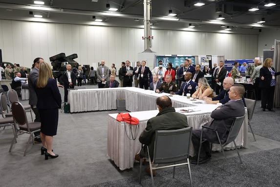 A large group of people sit around a white rectangular table in an exhibition hall. They are listening to two people (man and woman) in business attire speak.