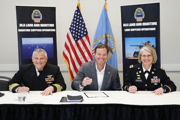 A man in a Navy Dress uniform, a man in a business suit and a woman in a Army dress uniform are seated at a white table and smile at the camera.