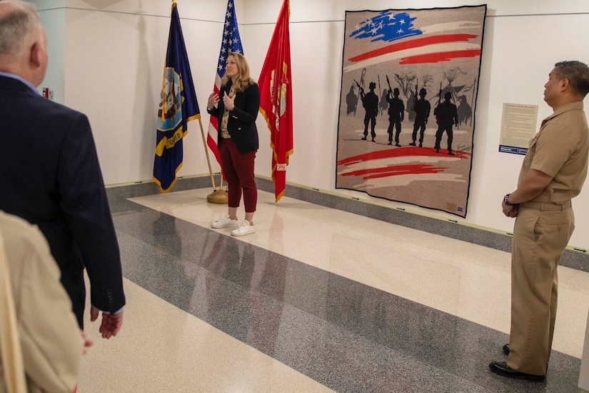 A civilian speaks by a blanket depicting U.S. troops that is displayed on a wall in a hallway where others stand listening.