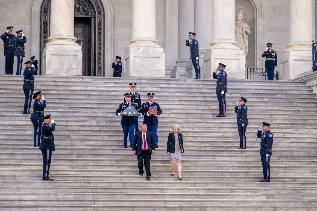 Service members stand in parallel formation on the U.S. Capitol steps as fellow service members carrying a folded flag and ashes walk down the center.