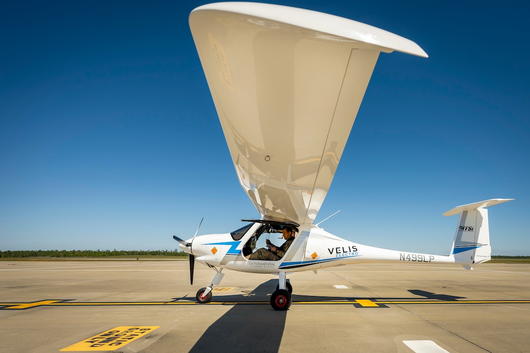 An airman sits in a small electric aircraft on a tarmac as see from under the wing.