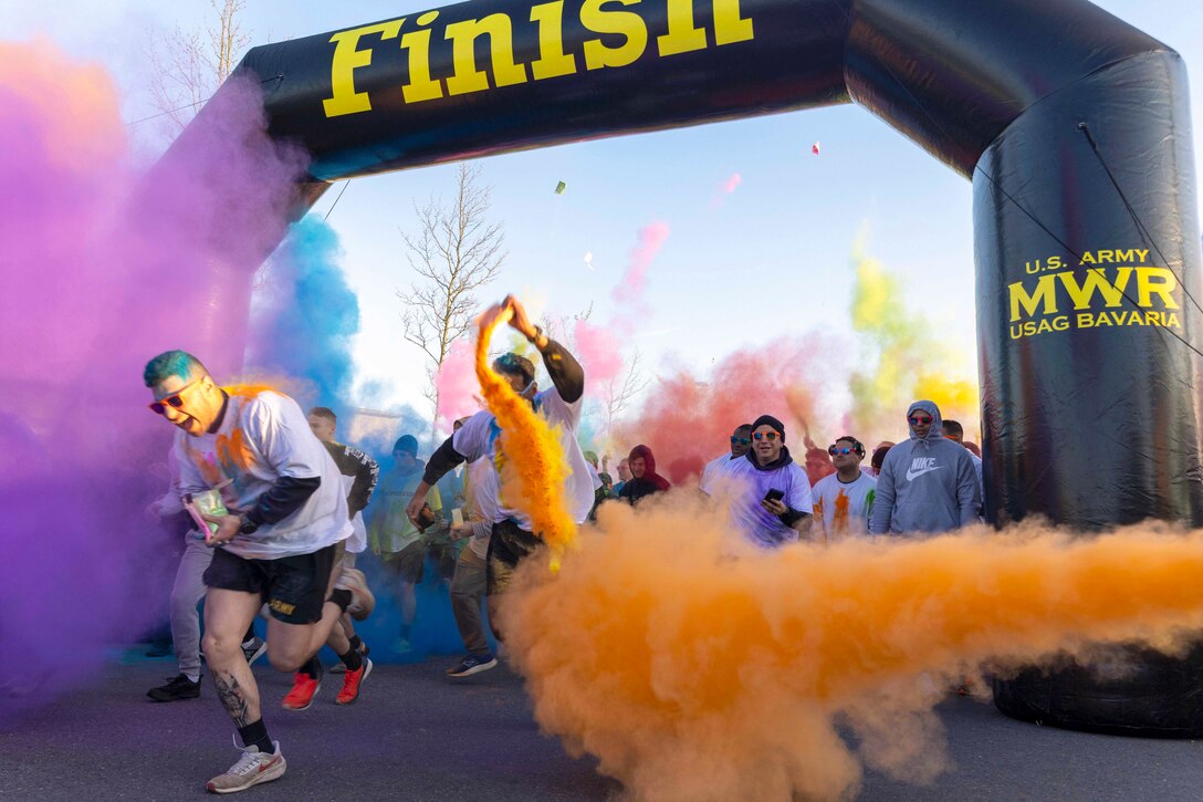 Dozens of participants run through the finish line of a race surrounded by colorful clouds of smoke.