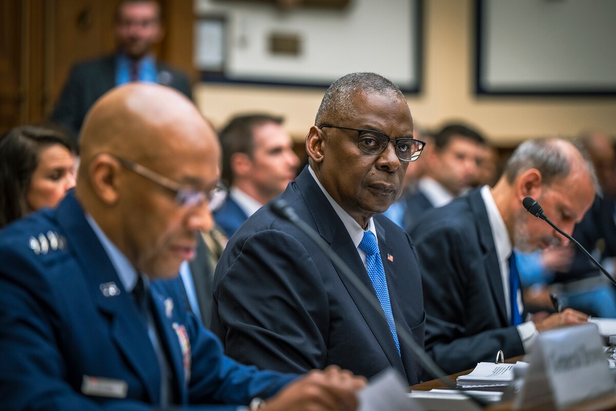Two men, one in a suit, one in a military uniform, sit near each other at a long wooden table.