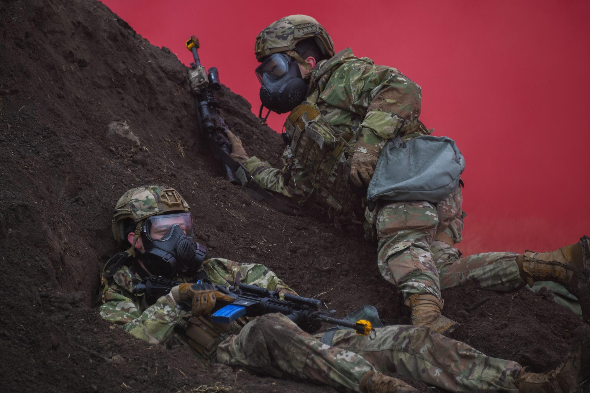 Two armed military members lay on dirt mound in smoke covered air.