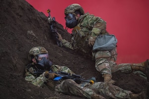 Two armed military members lay on dirt mound in smoke covered air.