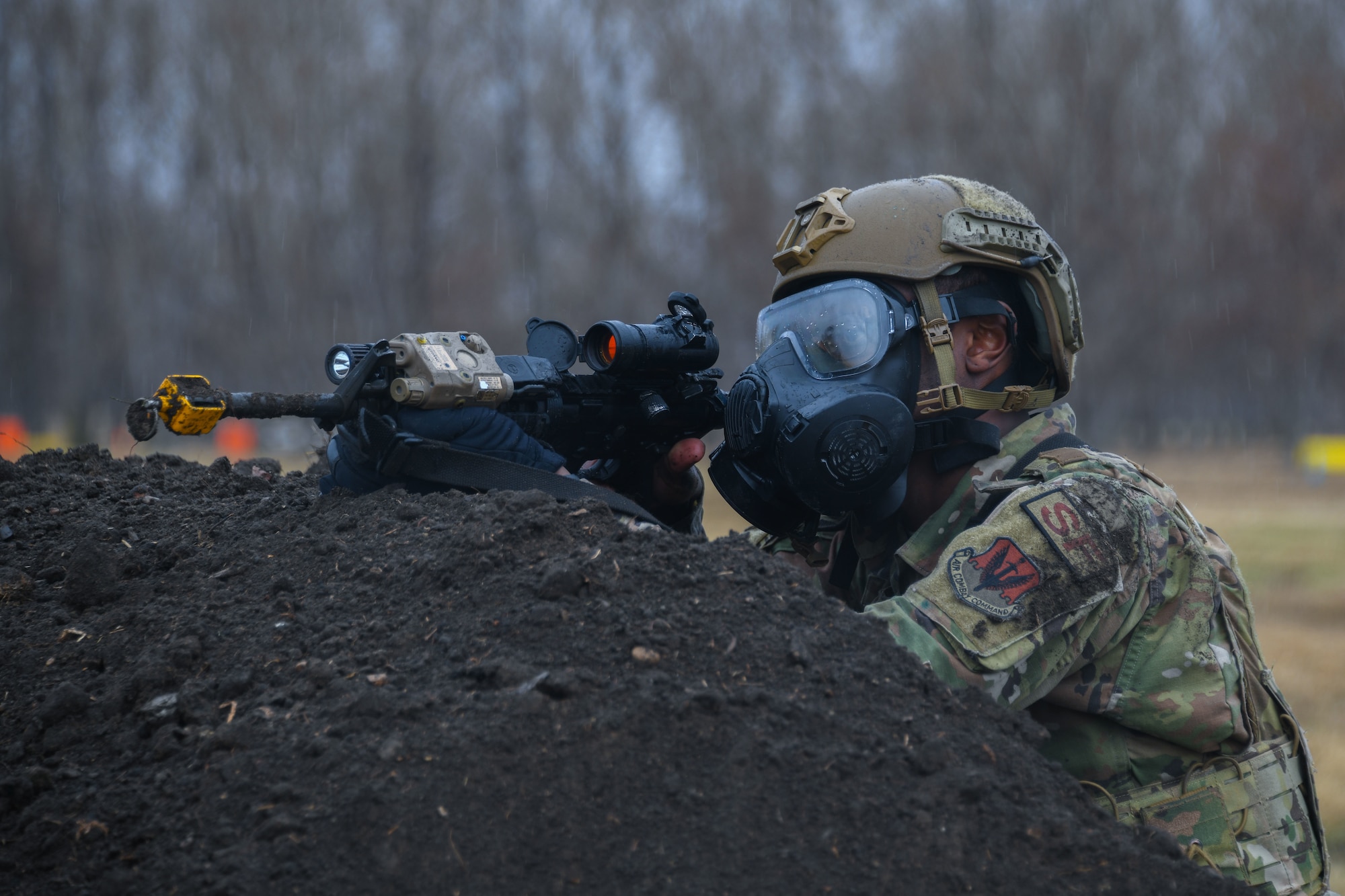 Armed military member with gas mask rests gun on top of dirt mound.