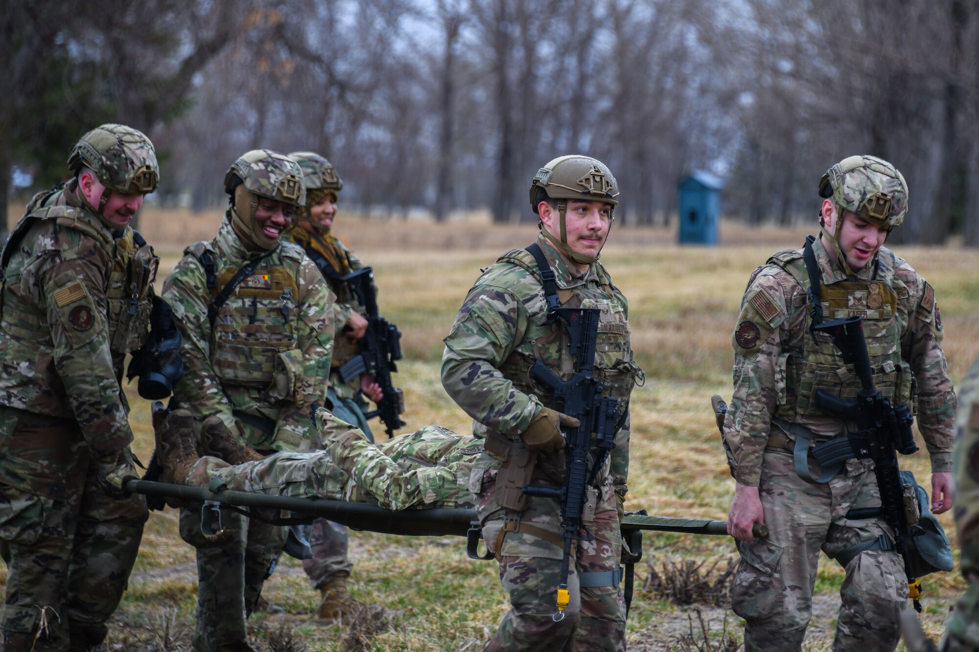 A group of military members carry an injured person on a stretcher in a field.