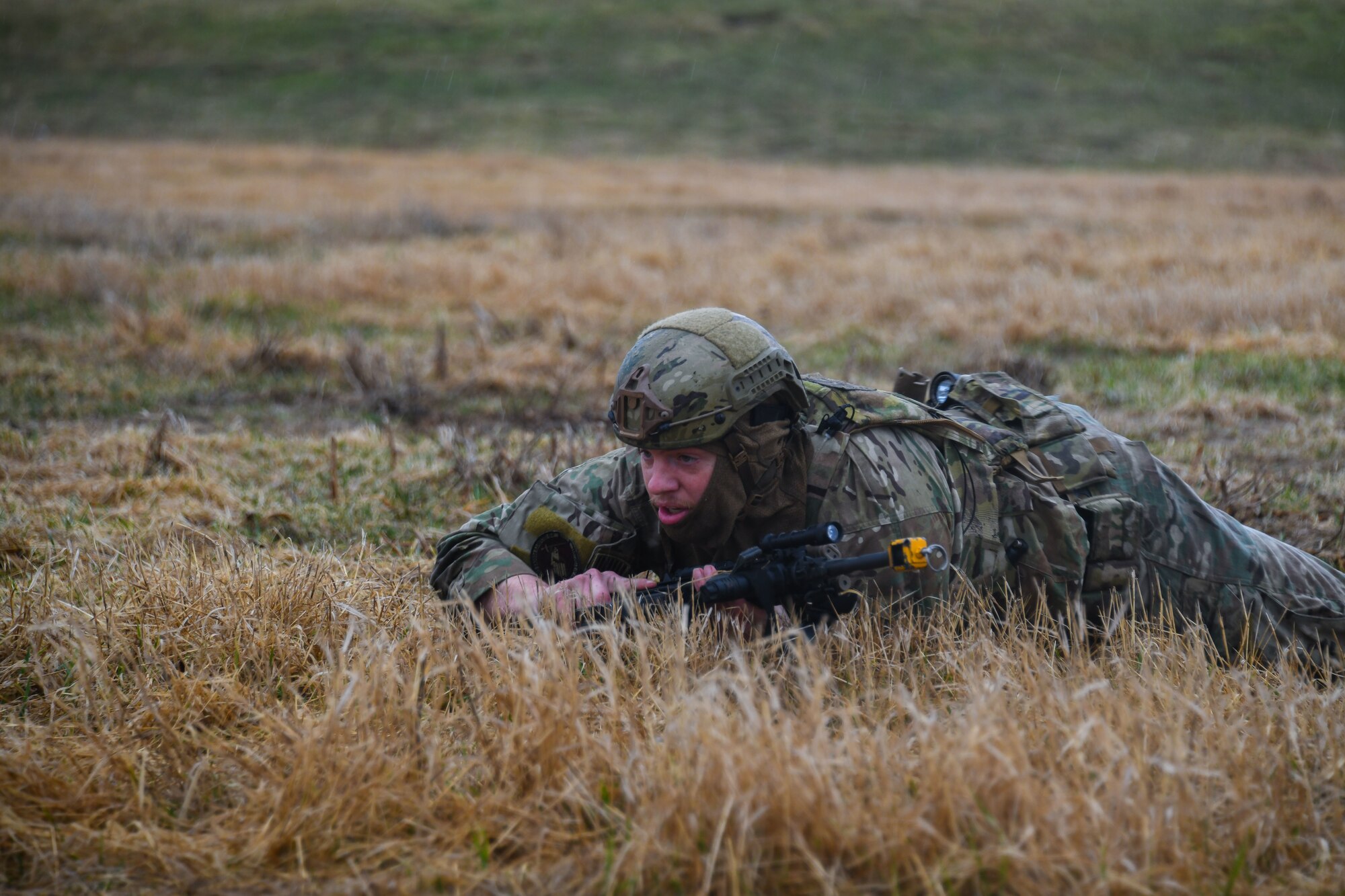 Military member low crawls in a field with a firearm.