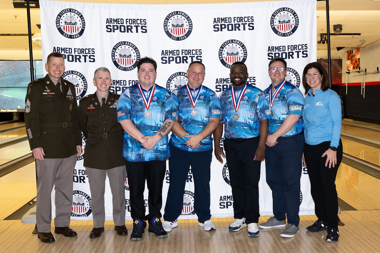 Medalists in a bowling tournament pose for a photo with uniformed service members in a bowling alley.