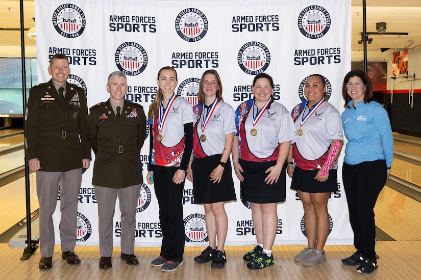 Medalists in a bowling tournament pose for a photo with uniformed service members in a bowling alley.