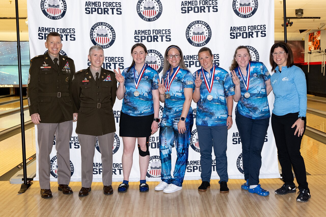 Medalists in a bowling tournament pose for a photo with uniformed service members in a bowling alley.