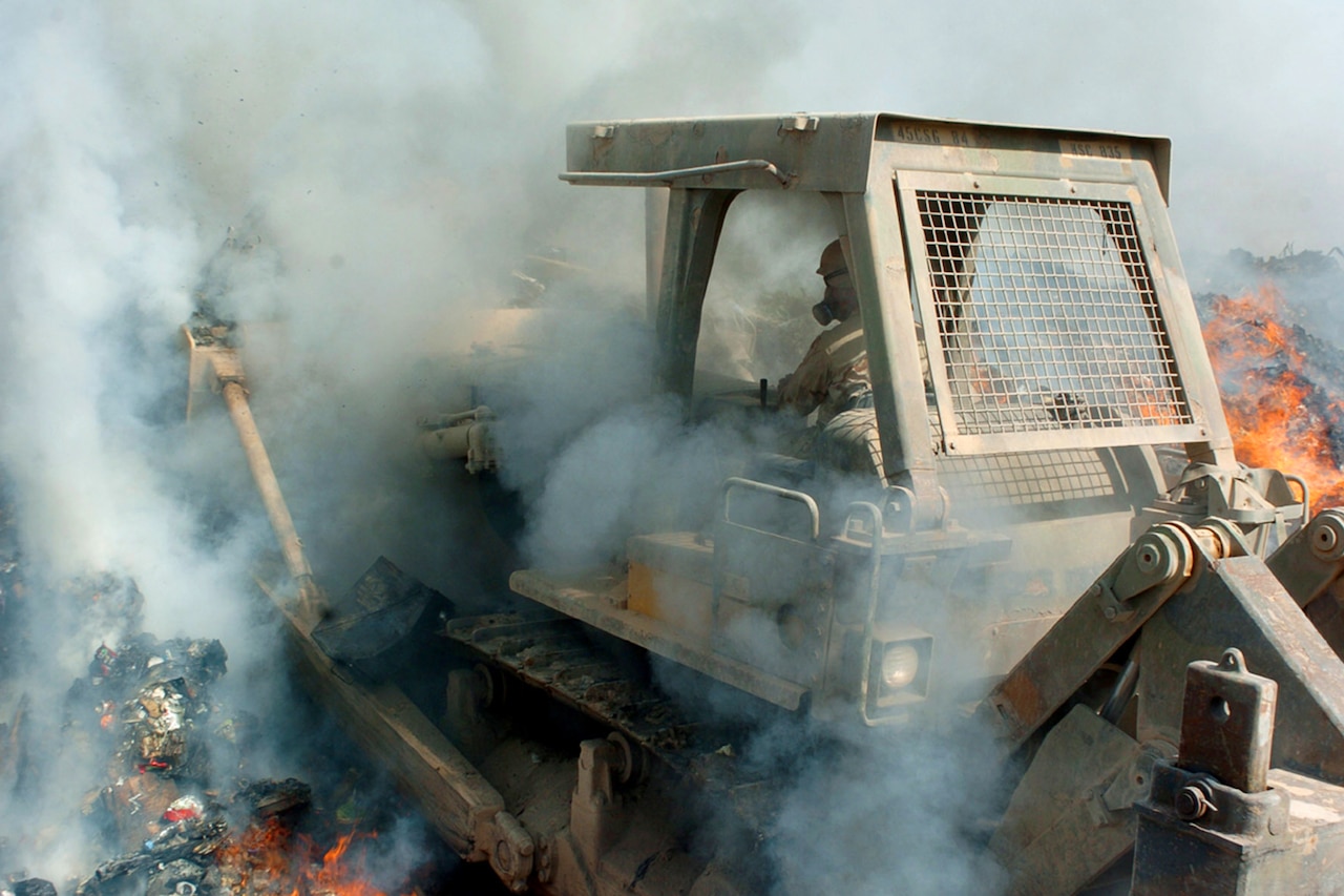 A bulldozer operator pushes garbage in midst of smoke and fire.