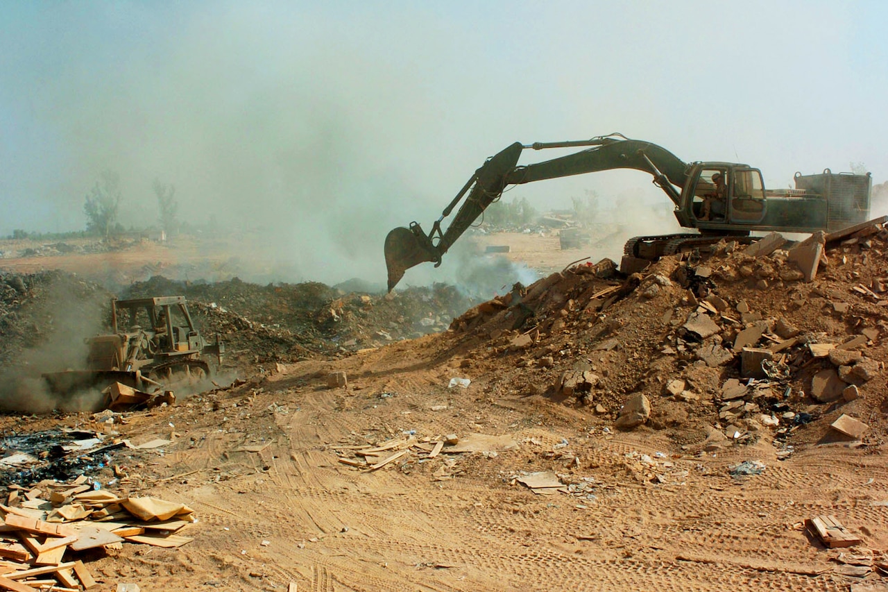 Bulldozer and excavator operators push garbage in midst of smoke.