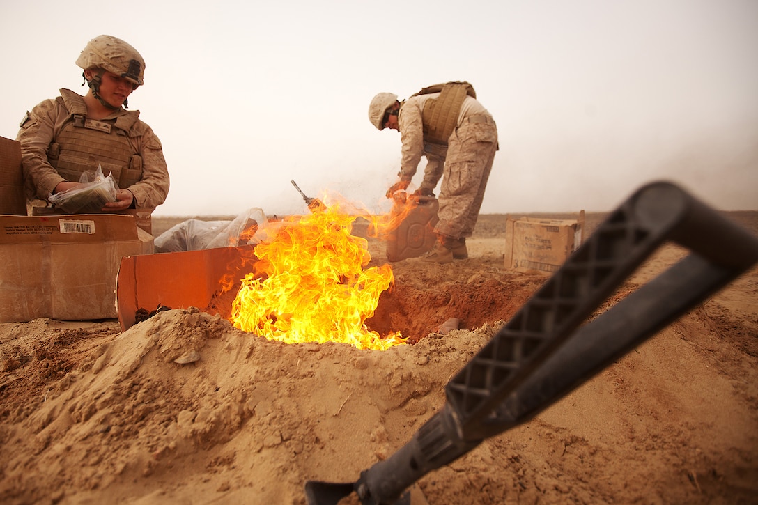 Two soldiers pick up trash from boxes next to a blazing fire pit in the desert.