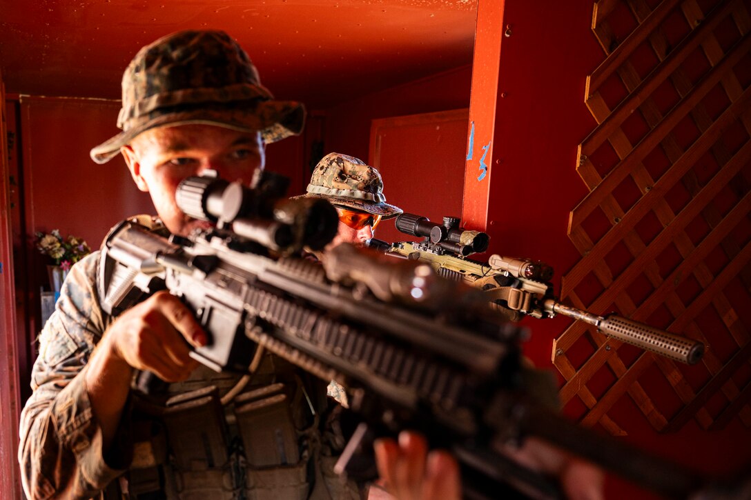 Two Marines point their weapons while looking ahead in a small red room.