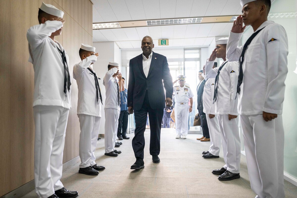 Secretary of Defense Lloyd J. Austin III walks between two rows of saluting sailors inside a building.