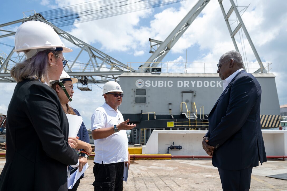 Three workers in business attire and hardhats speak with Defense Secretary Lloyd J. Austin III on a dock with Subic Drydock equipment in the background.