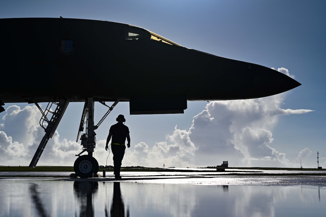 An airman walks under a parked aircraft with clouds in the distance as seen in silhouette.