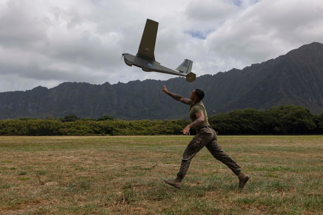 A Marine launches a small unmanned aerial system into the air while running in a field with mountains in the background.