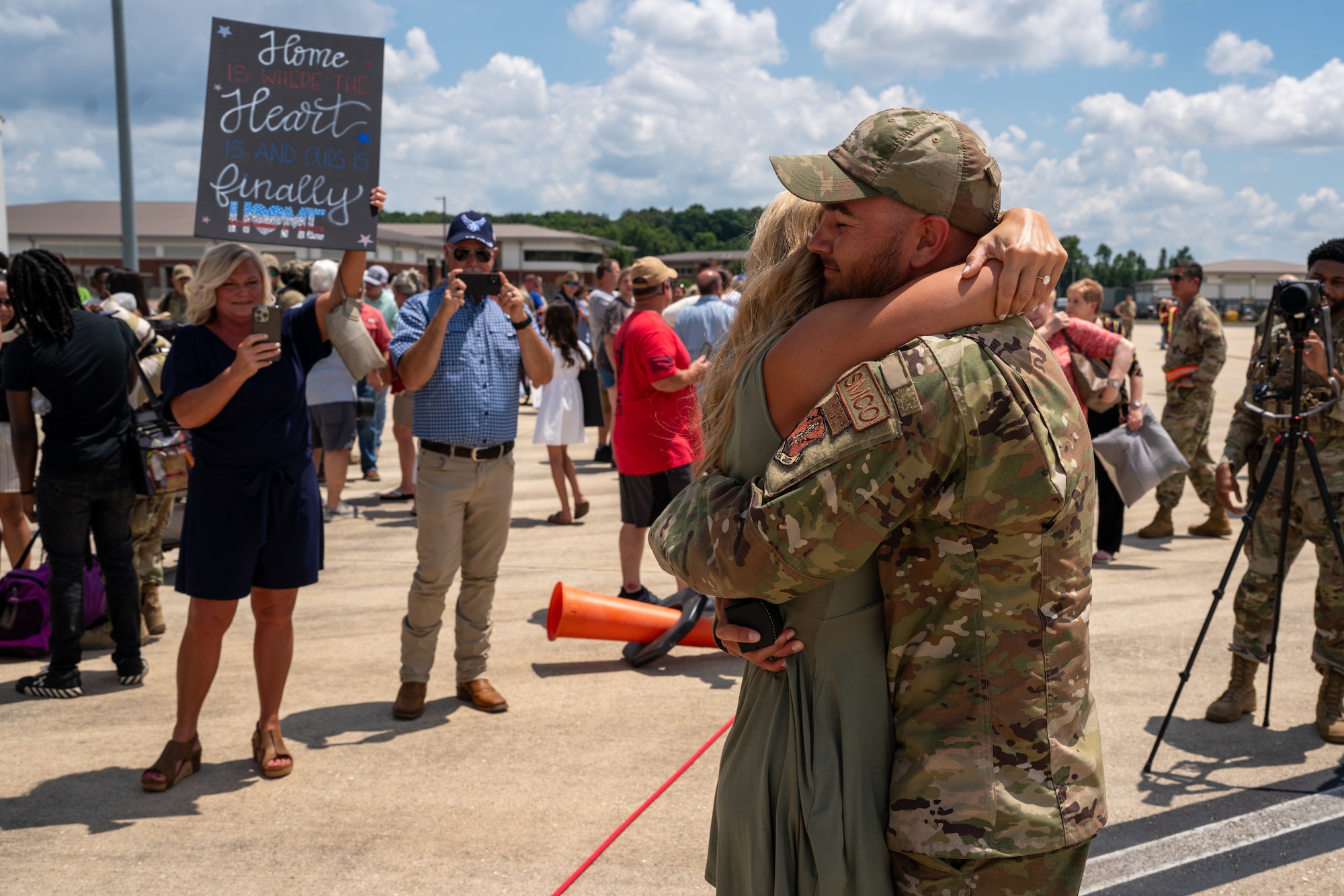 An Airman and a woman hugging in front of a crowd of people with a woman holding a sign in the background.