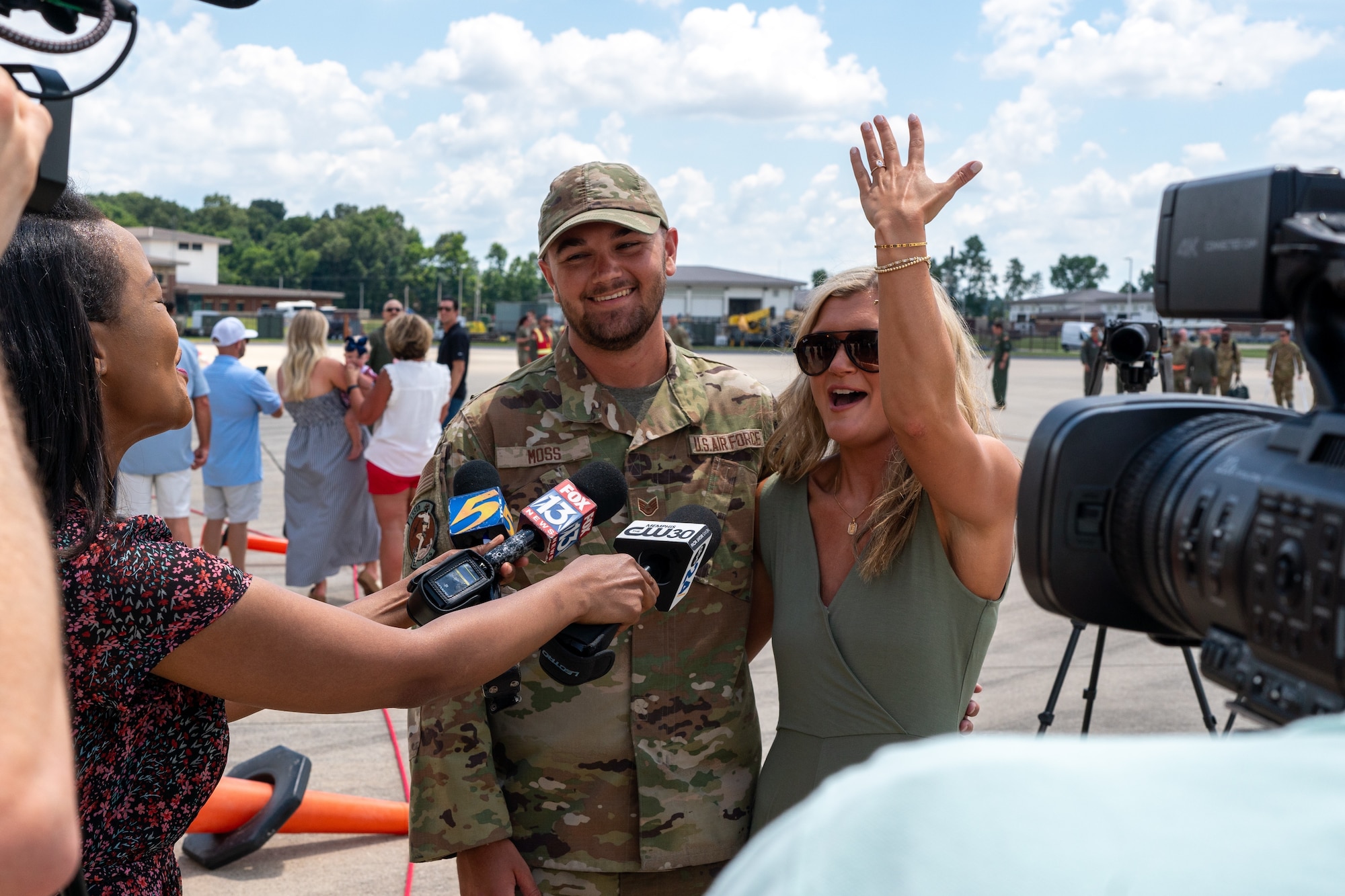 An Airmen and a young woman wearing green with her hand raised on a flight line with reporters pointing microphones at the them.