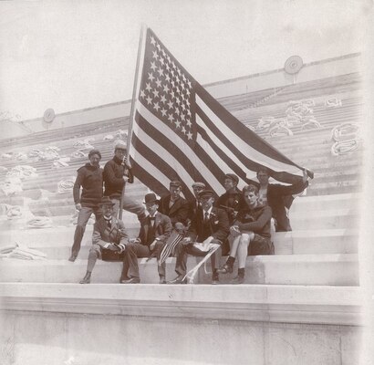 Group of men seated with two men standing, one of the men standing holds the American flag.