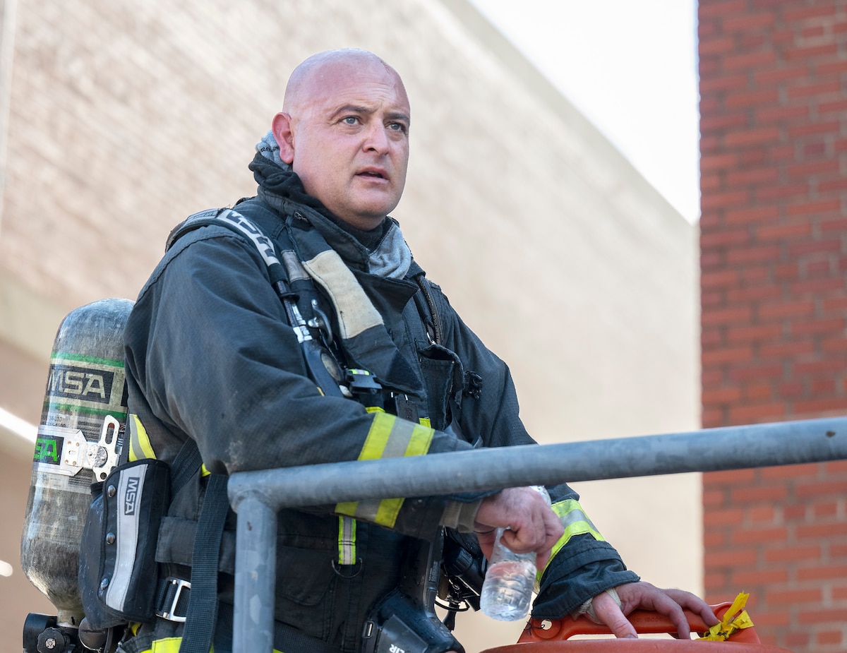 A bald firefighter, with an air tank on his back, leans against a handrail looking off into the distance with a bottle of water in his hand.