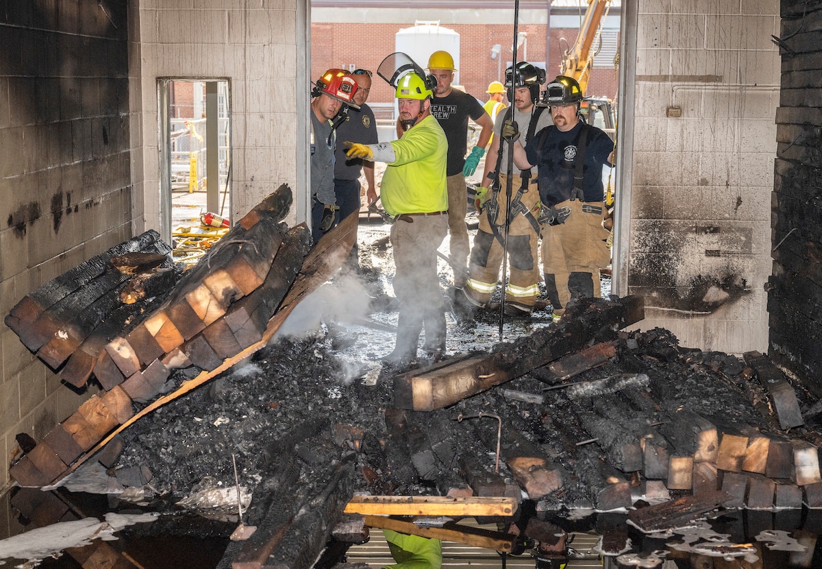A group of men in firefighter helmets and hardhats stand looking at burnt wood in a room.