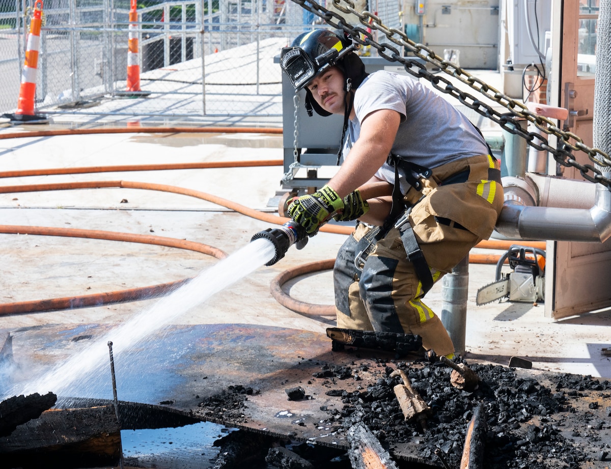 A firefighter stands with bent knees, in fireman pants and a t-shirt using a hose to spay charred items outside a building.