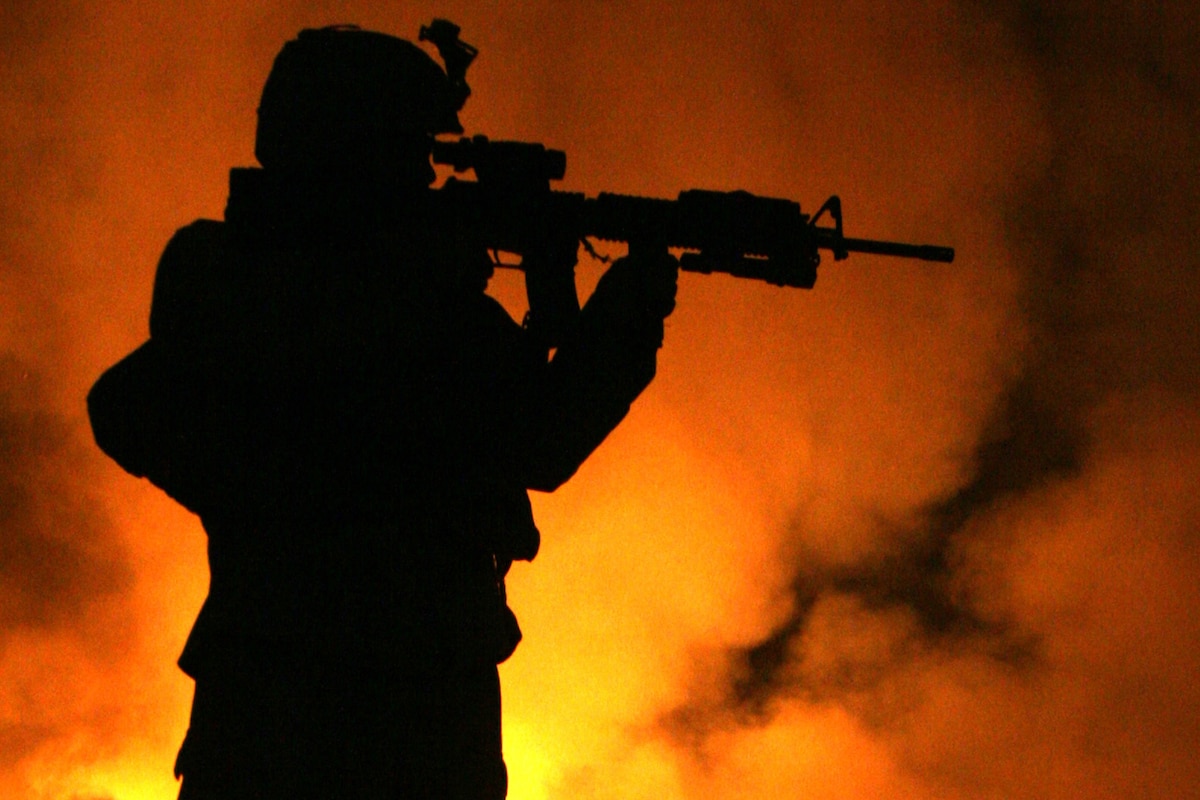 Fire rages behind a standing Marine with a rifle raised in firing position.