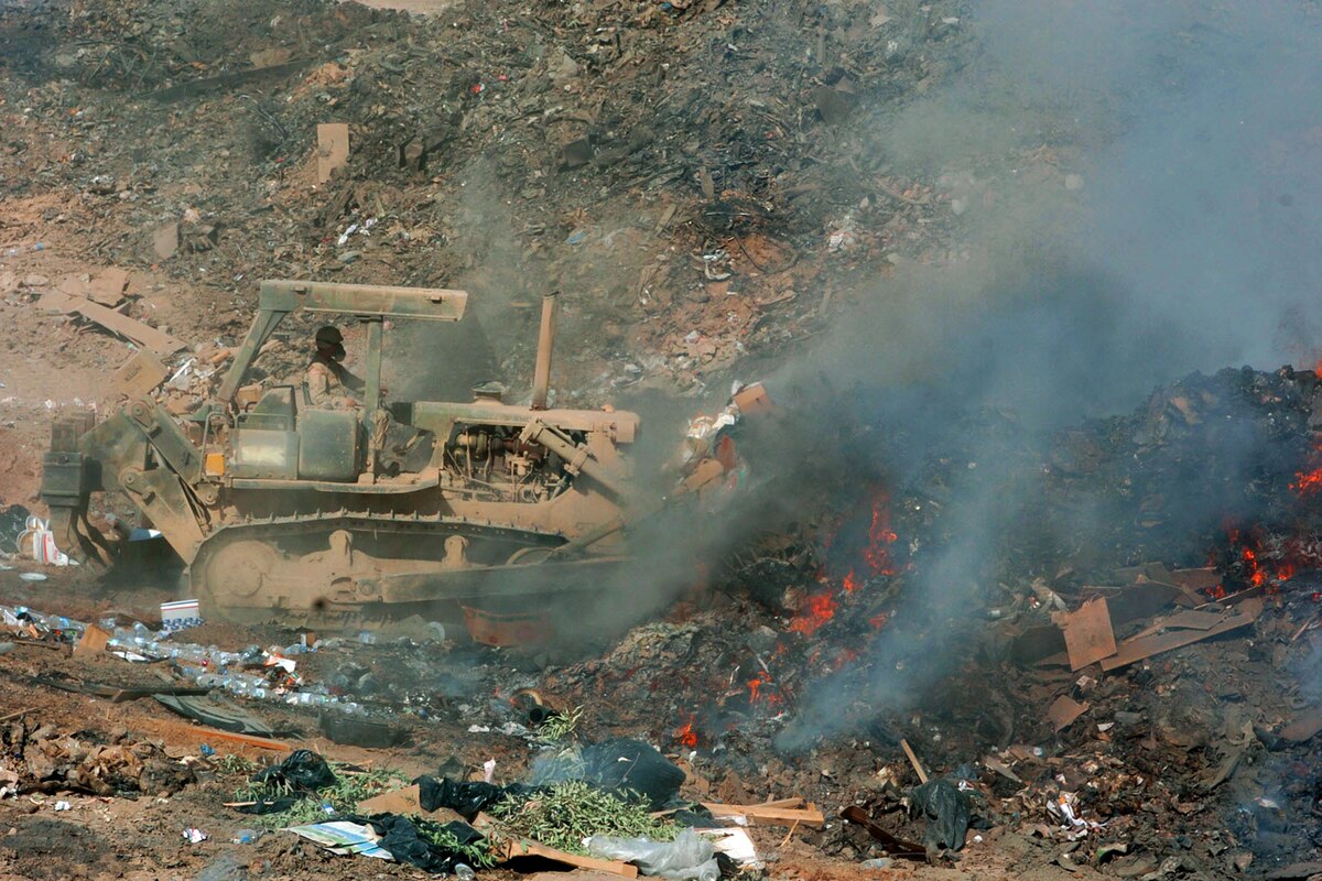 A bulldozer operator pushes garbage in midst of smoke and fire.