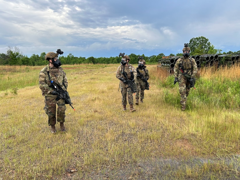 Four soldiers in tactical gear and masks walk through a grassy field, with military equipment visible in the background under a cloudy sky.