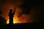 A soldier stands silhouetted in black in front of a fire pit at night.