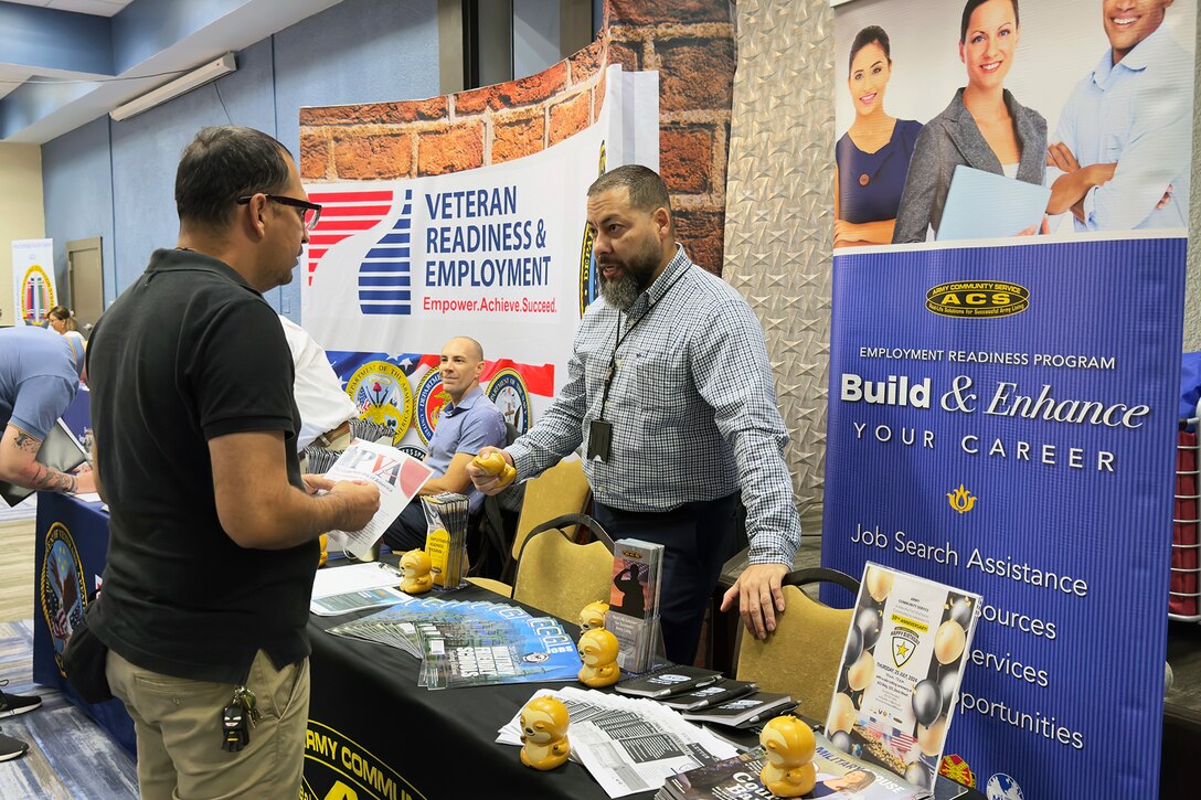 Two civilians talk on either side of a table display, with posters featuring career-related messages hanging behind them.