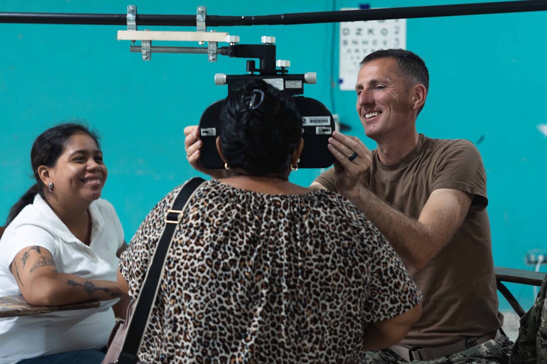 A Navy officer smiles while giving a patient an eye exam. A second person smiles and watches.