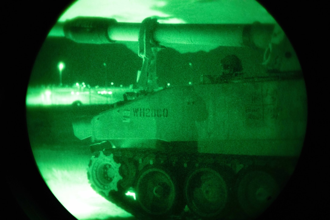 A green night vision photo seen through a scope shows a soldier sitting in a howitzer with lights in the distance.