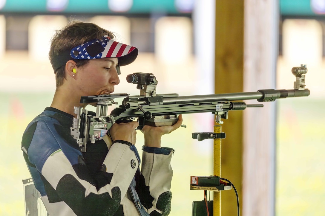 A soldier wearing an athletic uniform stands and looks through the scope of a marksman’s rifle attached to a thin pedestal.