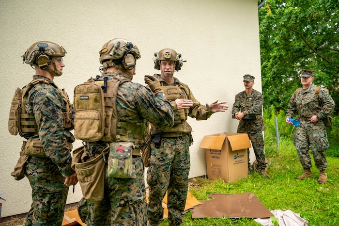 Two U.S. Marines in helmets and vests receive instruction from another Marine in a helmet and vest outside of a building.