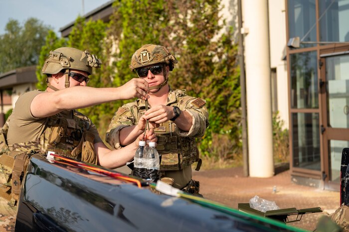Two U.S. Air Force Airmen in helmets and vest slide a wire into two taped-together water bottles standing upright on the tailgate of a truck.