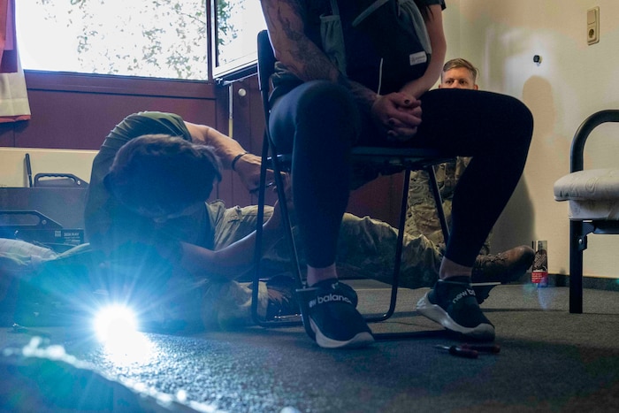 A Lithuanian Army Soldiers under a chair occupied by a civillian in an office. A flashlight by the chair shines right into the camera and a U.S. Air Force Airmen watches from a corner behind the chair.