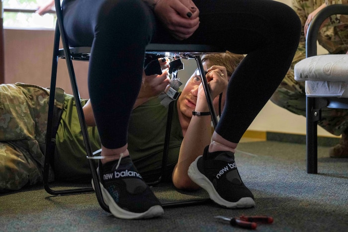 A German Army solider shines a flashlight and looks under a chair occupied by a person in civilian clothes in an office