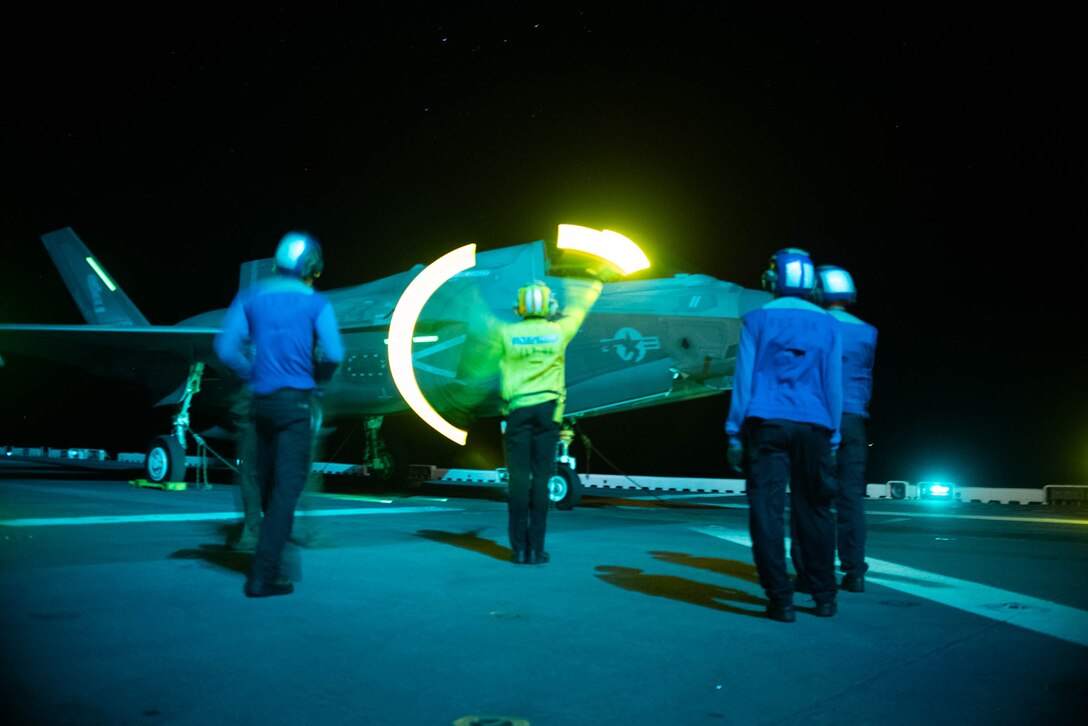 A sailor uses air traffic control lights to direct a military aircraft on a ship's flight deck at night as others watch.