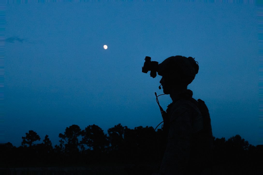 Silhouette of a service member at twilight wearing a helmet with the moon and a tree line in the distance.