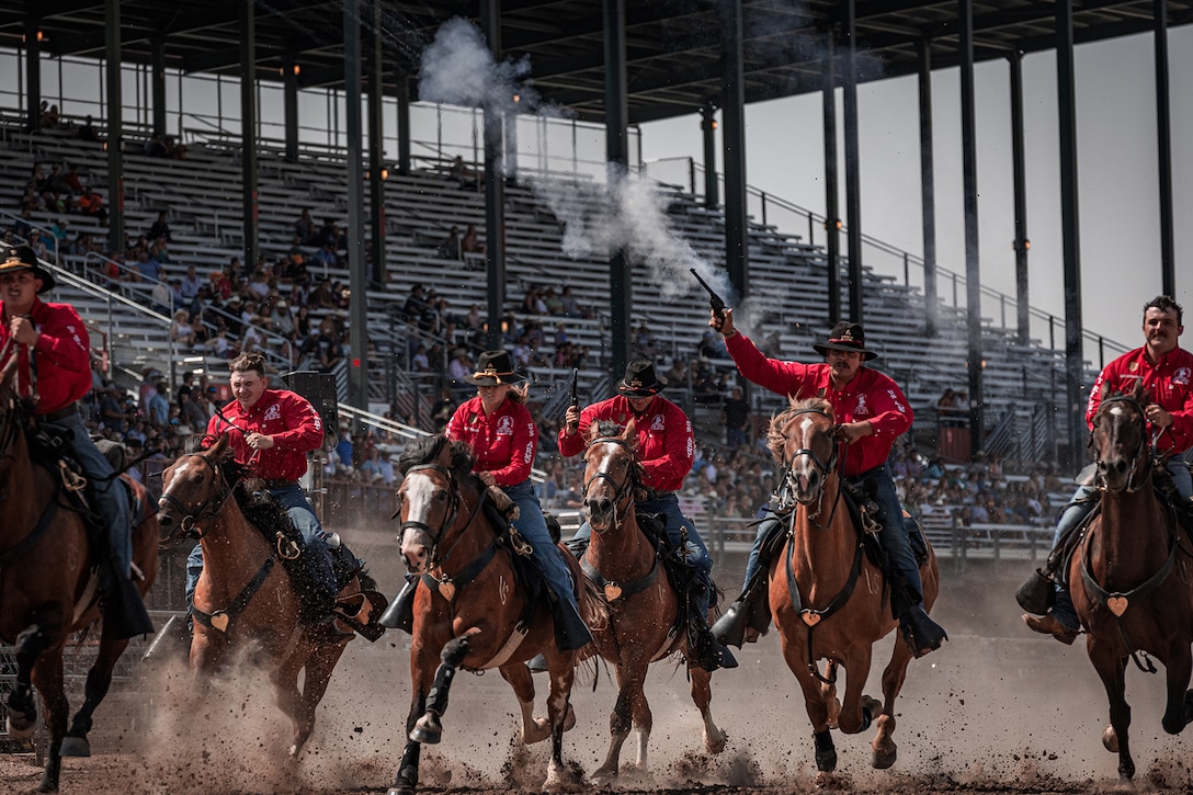 Service members ride horses, kicking up dust, as people watch from the stands. One soldier fires a weapon while on horseback.