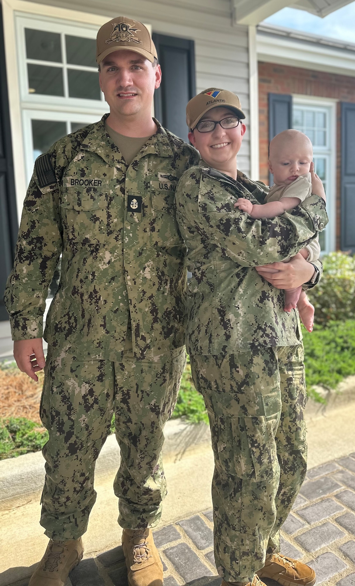 Two Navy personnel pose as partners in front of their home with their child outside of their home.
