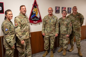 Five soldiers pose in front of a military wall emblem.