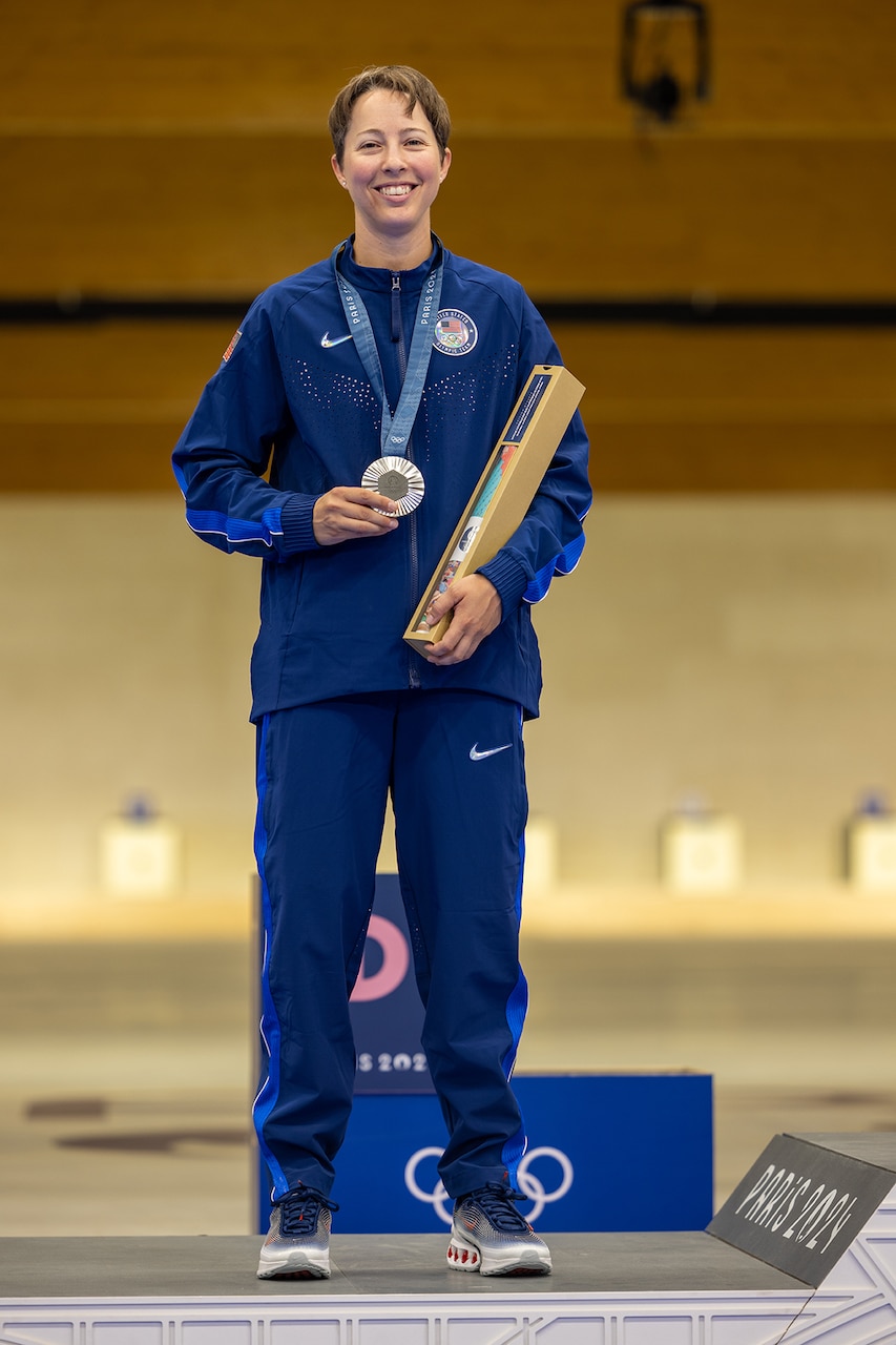 A soldier in athletic wear and a silver medal stands on a podium while holding a box.