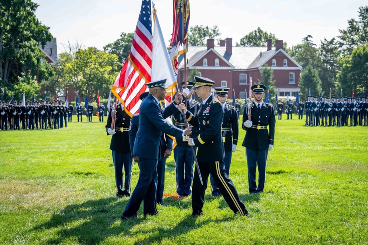 Two people in military dress hold a flag in a grass field with other service members standing in formation in the background,